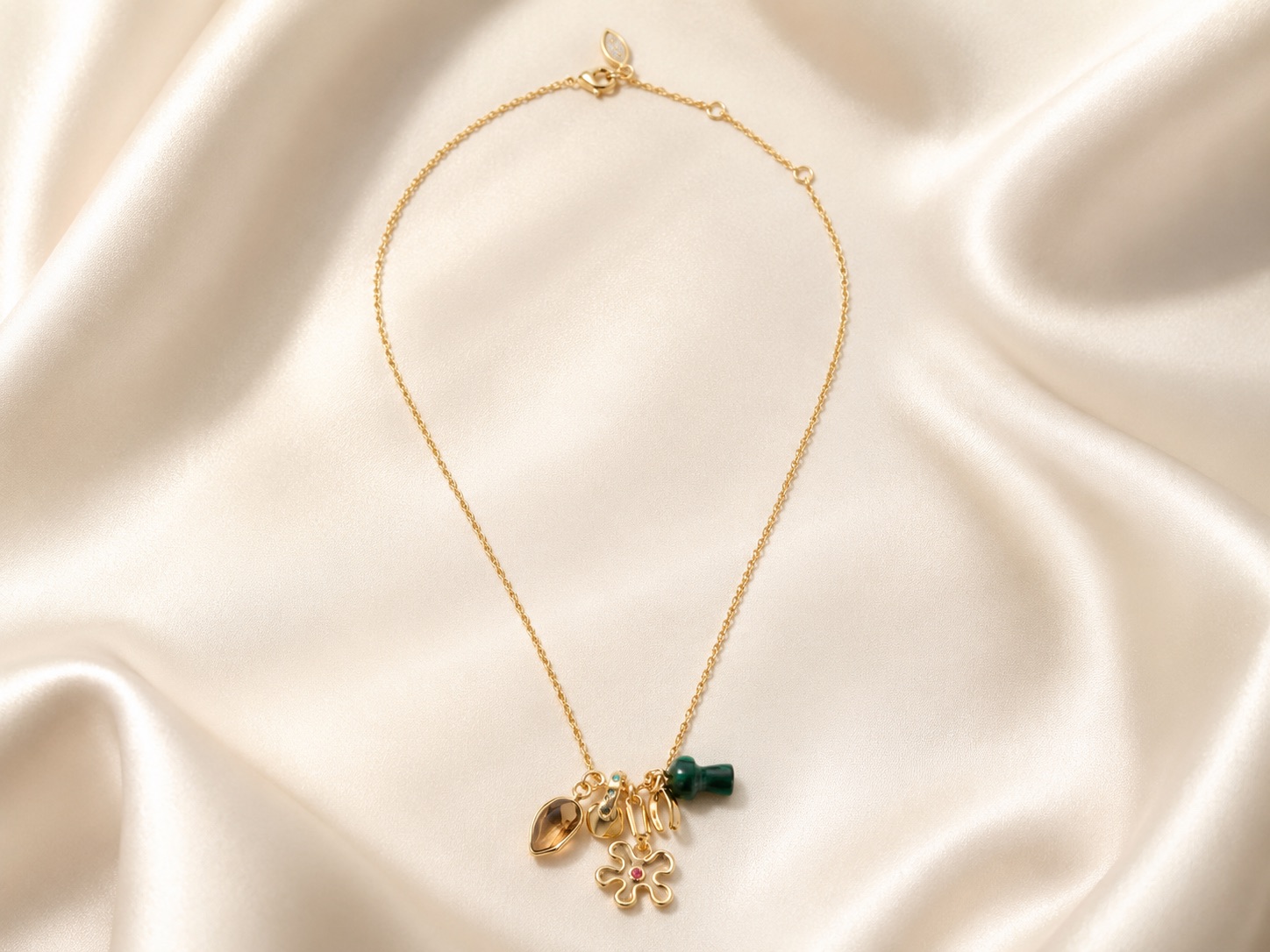 Goldfarbene Kette mit unterschiedlichen Anhängern, darunter grüner Stein, florales Motiv und kleiner Ring